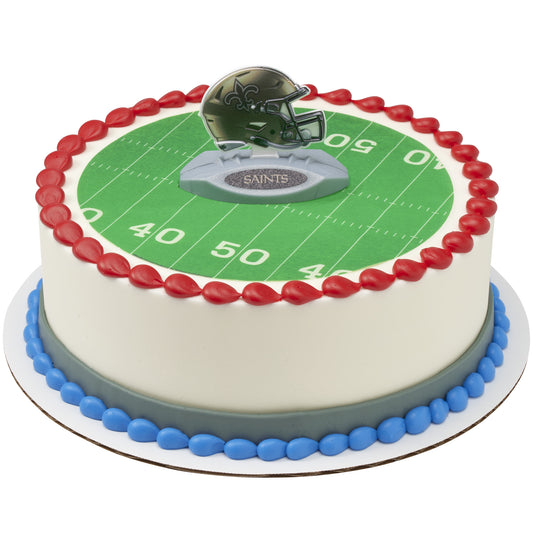 A round cake decorated like a football field, with a green yard-line top surface, red piped border, blue base trim, and a helmet topper on a stand. The side of the cake is white, and the word 'SAINTS' is visible on the helmet topper.