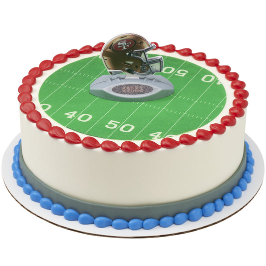A round cake decorated with a football field design, red piped border, blue base trim, and a small helmet topper on top, shown on a white background.