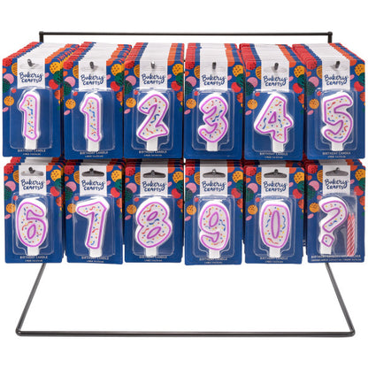 Display rack holding multiple packaged numeral birthday candles (0–9) with purple outlines and confetti centers on blue Bakery Crafts backing cards.