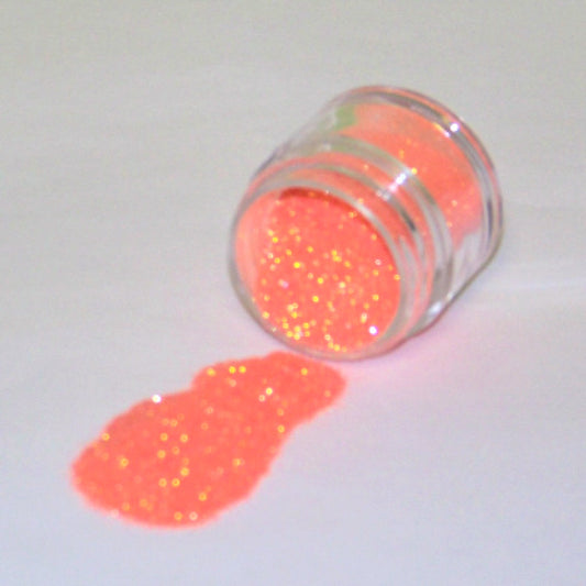 Small clear jar tipped on its side spilling bright orange glitter dust onto a white surface, showing fine sparkling particles with a vivid neon orange glow.