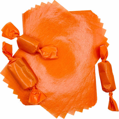 Three rectangular caramel pieces individually packaged in bright orange wax candy wrappers with twisted ends, arranged around a stack of 4" x 5" caramel wrapper sheets.