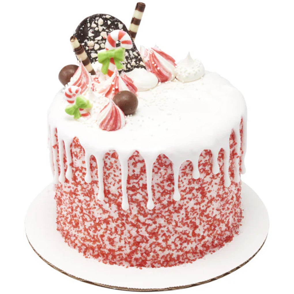 White drip cake on a round cake board with red and clear peppermint sanding sugar pressed around the sides, topped with candy decorations.