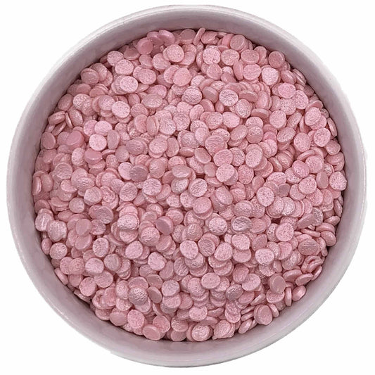 Top-down view of a bowl filled with pearlized pink confetti sprinkles, featuring small round edible discs with a soft shimmer finish.