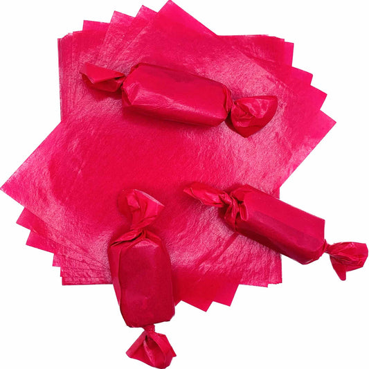 Three rectangular caramel pieces individually packaged in vivid pink wax candy wrappers with twisted ends, arranged over a stack of 4" x 5" caramel wrapper sheets.