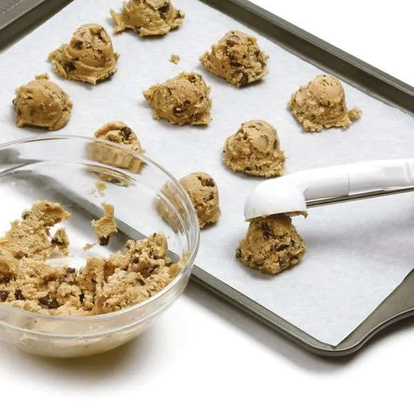 Cookie dough scoop with white handles placing dough balls onto a parchment-lined baking sheet.