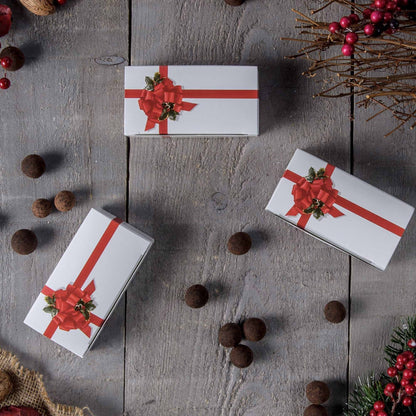 Red ribbon and holly candy boxes styled on a rustic wood surface with scattered chocolate truffles and holiday greenery accents.