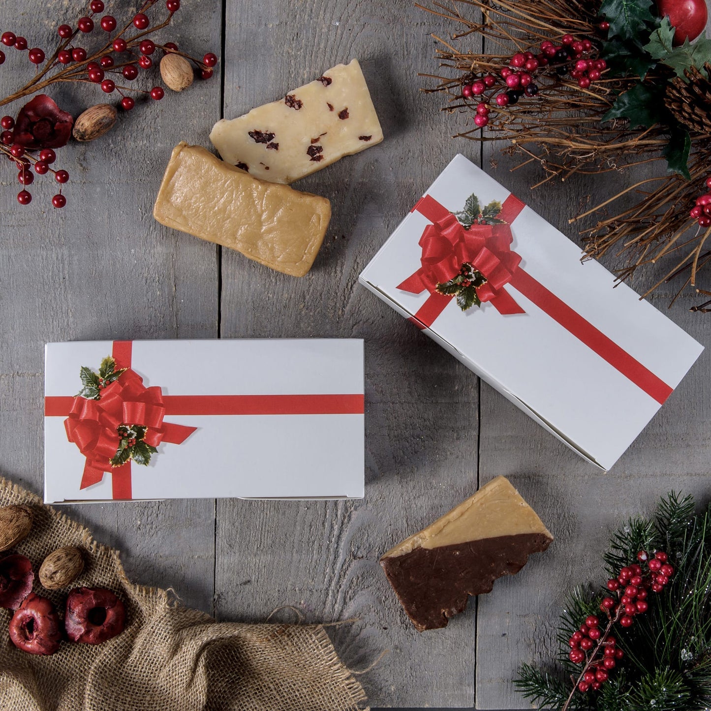 Holiday gift boxes with red ribbon and holly bow design styled on a rustic wood surface with fudge pieces and seasonal greenery and berries.