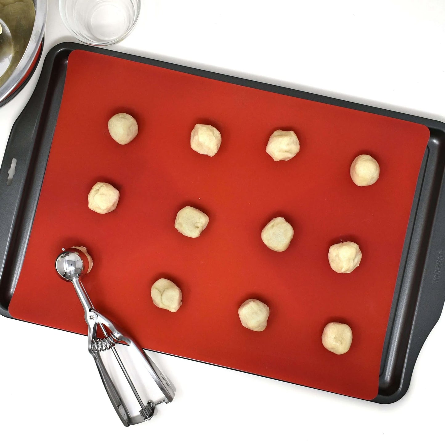 Red silicone baking mat placed on a dark baking sheet with small dough balls arranged in rows and a metal cookie scoop nearby.
