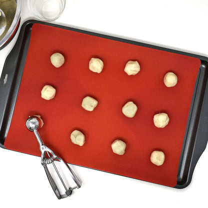 Red silicone baking mat placed on a dark baking sheet with small dough balls arranged in rows and a metal cookie scoop nearby.