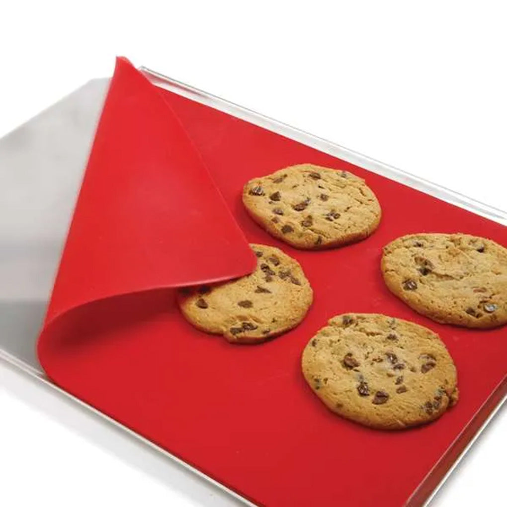 Red silicone baking mat on a metal baking sheet with several baked chocolate chip cookies, with one corner of the mat lifted to show the flexible texture.