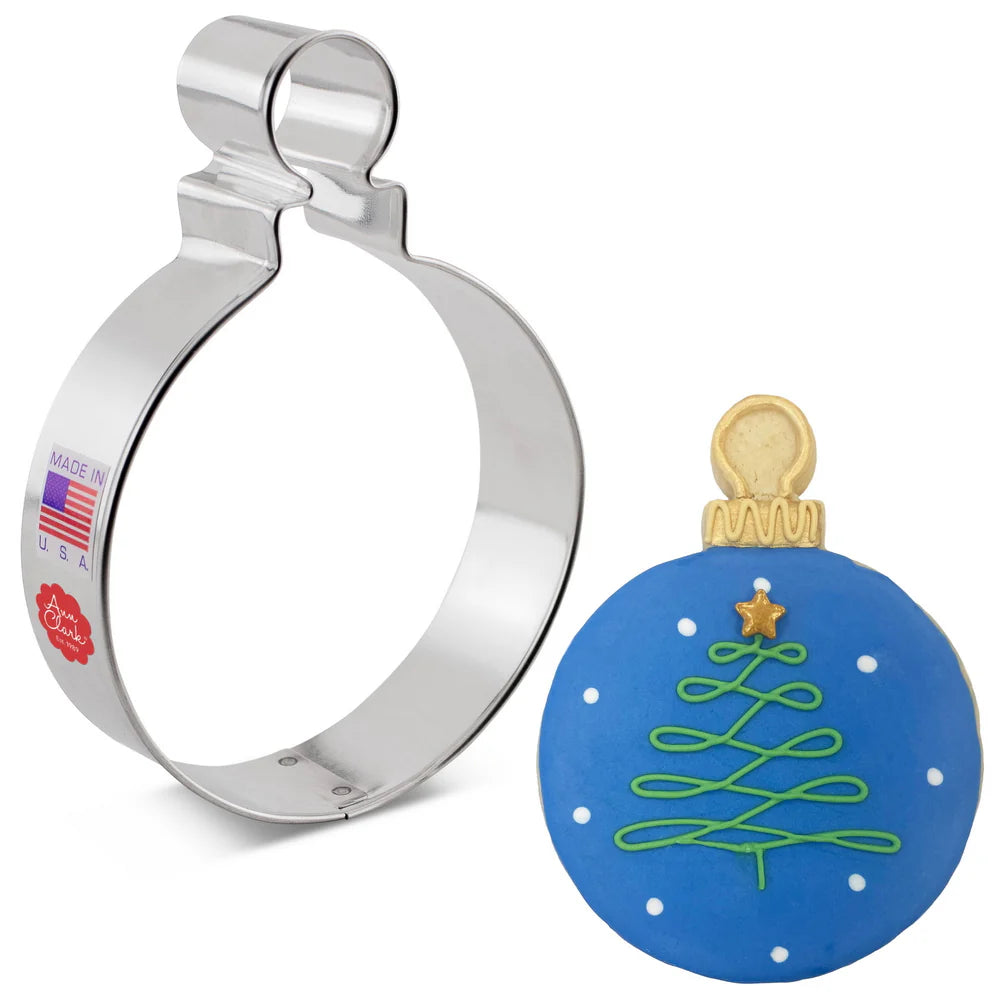 Round Christmas ornament cookie cutter next to a decorated cookie with blue icing, a gold ornament top, and a green piped tree design.