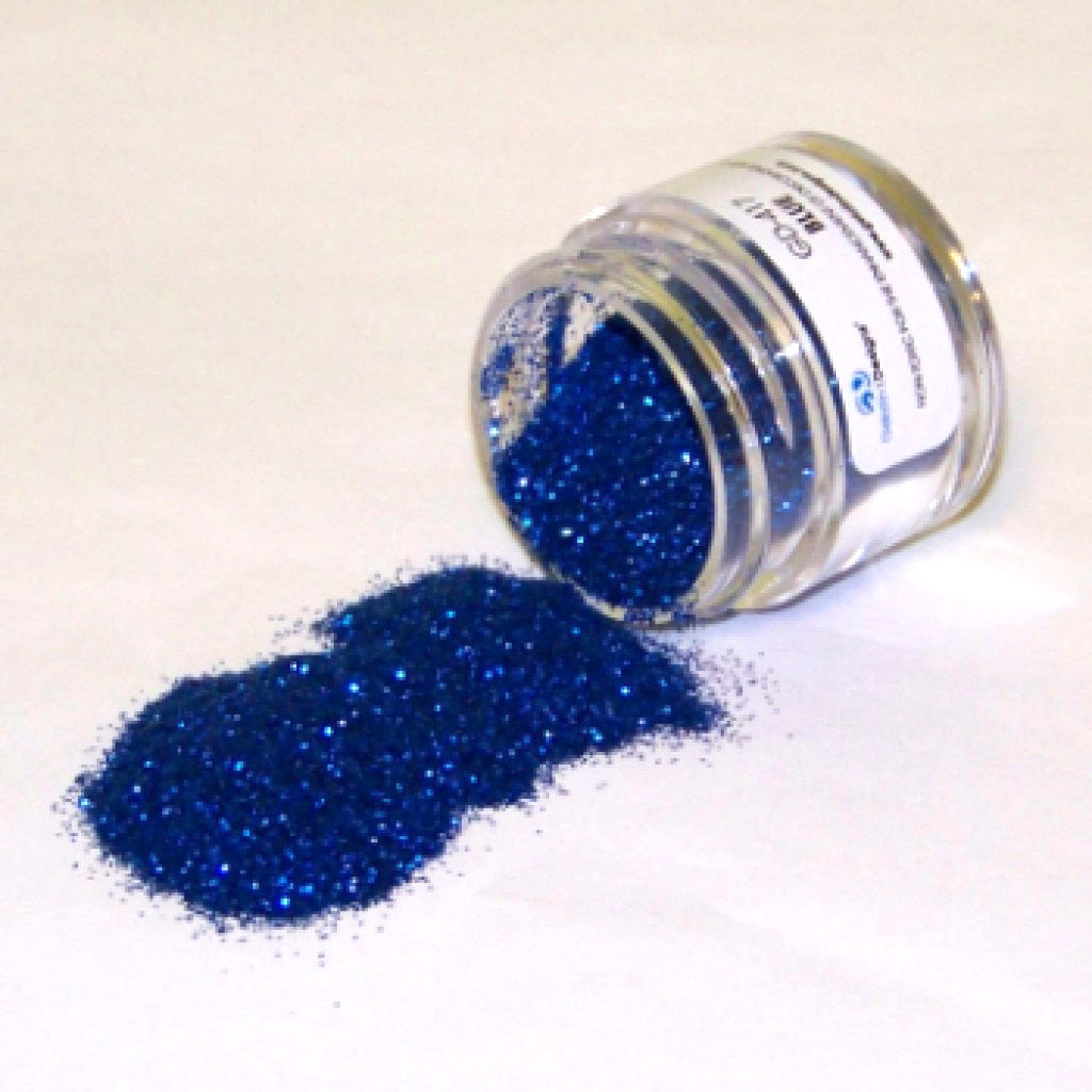 Small clear jar tipped on its side spilling royal blue glitter dust onto a white surface, showing dense sparkling blue particles.