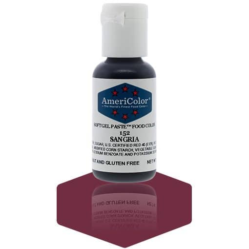 A bottle of AmeriColor Sangria soft gel paste food coloring with a white cap, blue label, and red stars, standing on a rich sangria-red reflective surface that mirrors the bottle.