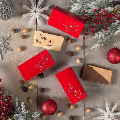 Holiday flat-lay with red Season’s Greetings candy boxes on a rustic wood surface, surrounded by ornaments, frosted greenery, berries, and assorted candy pieces.