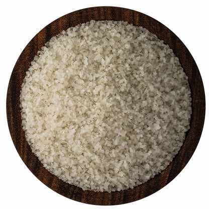 Overhead view of fine French grey sea salt crystals in a round dark wooden bowl on a white background, showing pale grey-beige grains.