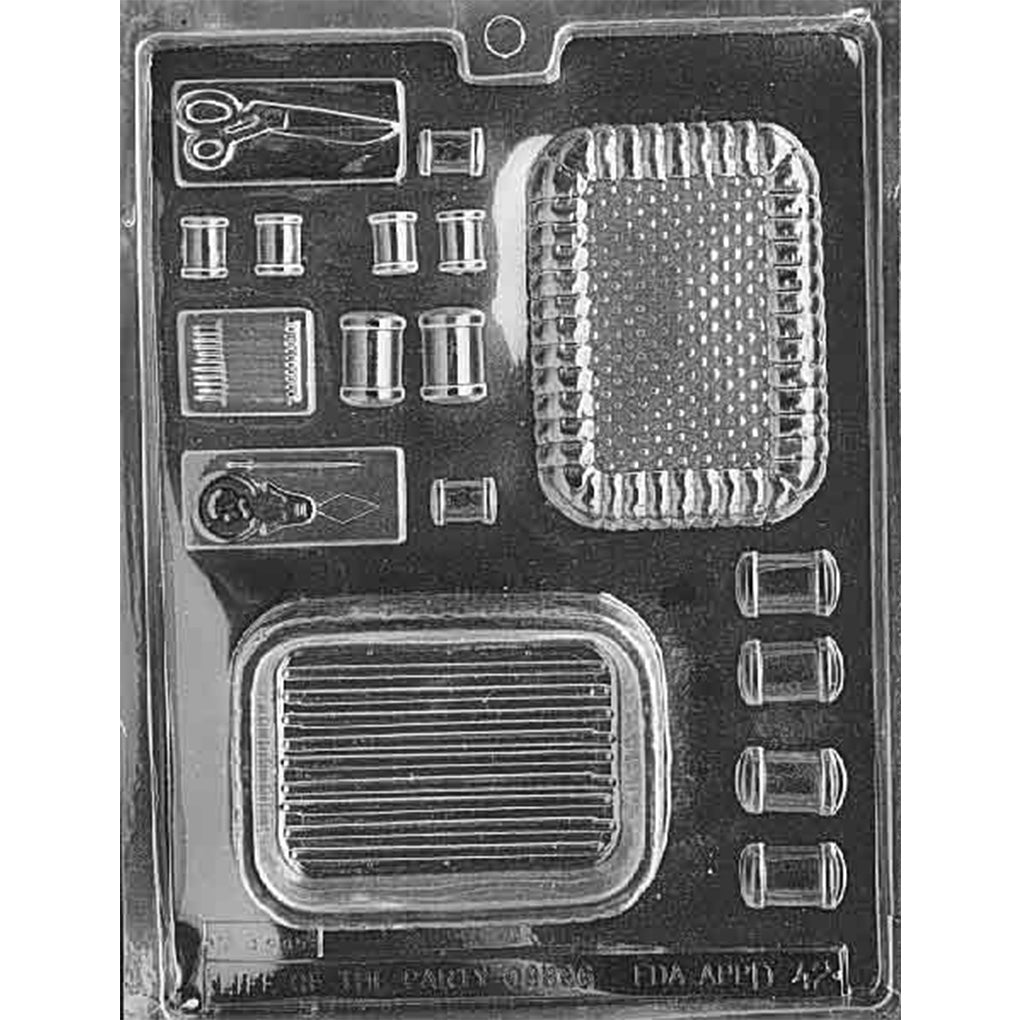 Clear plastic chocolate pour box mold with sewing kit shapes including thread spools, scissors, buttons, and a rectangular box lid and base, shown on a dark background.