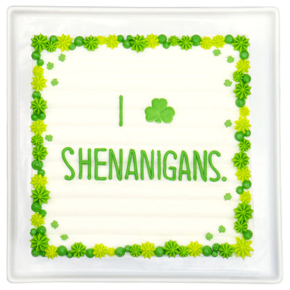 Rectangular sheet cake with a green piped border and small shamrock accents, featuring the word “SHENANIGANS” in green icing on a white background.