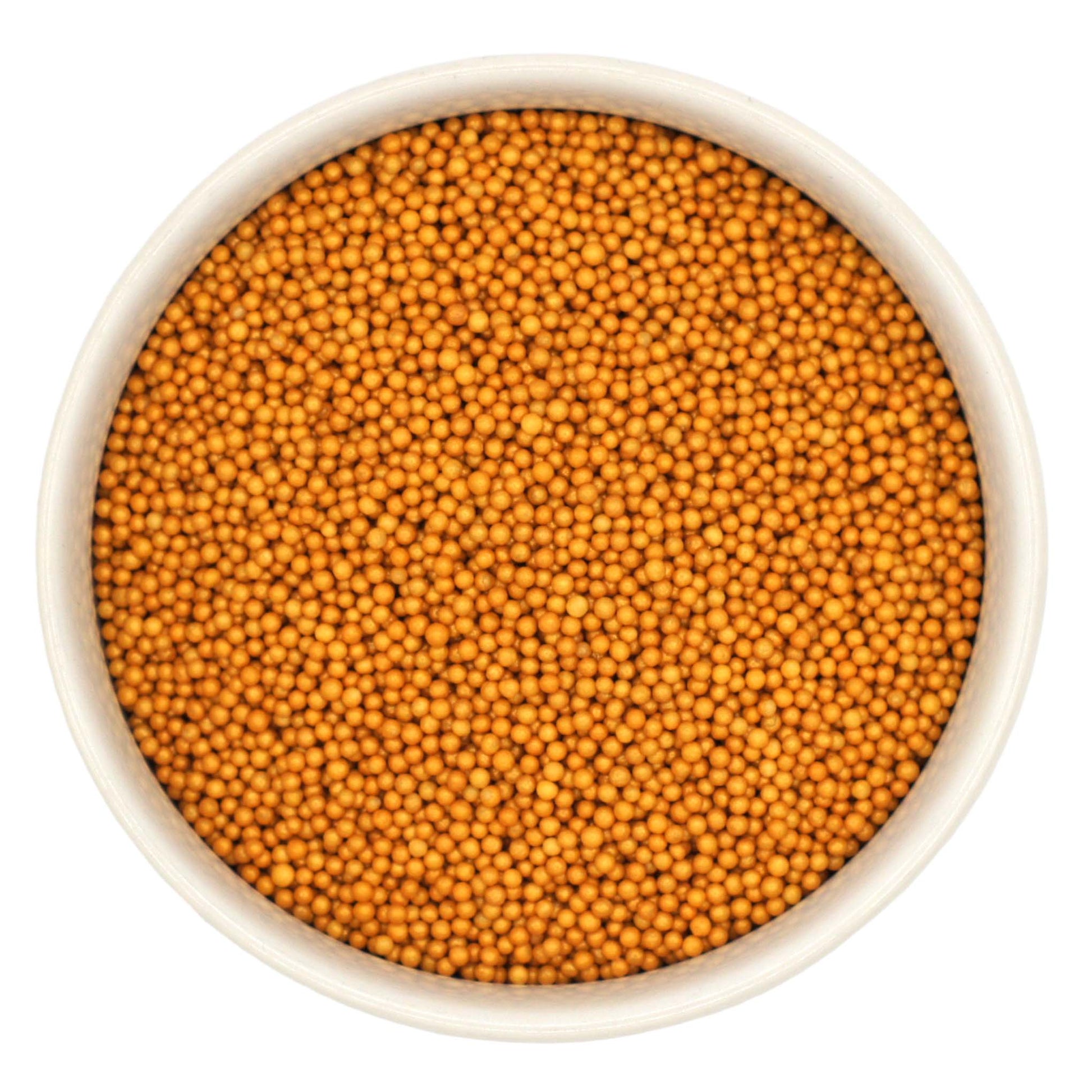 Top-down view of a white bowl filled with shimmer gold non-pareils, showing tiny round metallic sprinkle beads in a rich golden yellow shade.