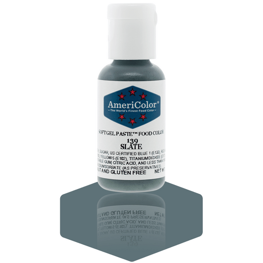 A bottle of AmeriColor Slate soft gel paste food coloring with a white cap, blue label, and red stars, standing on a dark slate-gray reflective surface that mirrors the bottle.