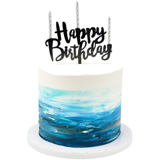 White and blue watercolor-style frosted cake topped with three silver spiral candles and a black “Happy Birthday” topper.