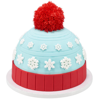 Winter hat-themed cake decorated with multiple white pressed sugar snowflakes on blue fondant and a large red pom-pom on top.