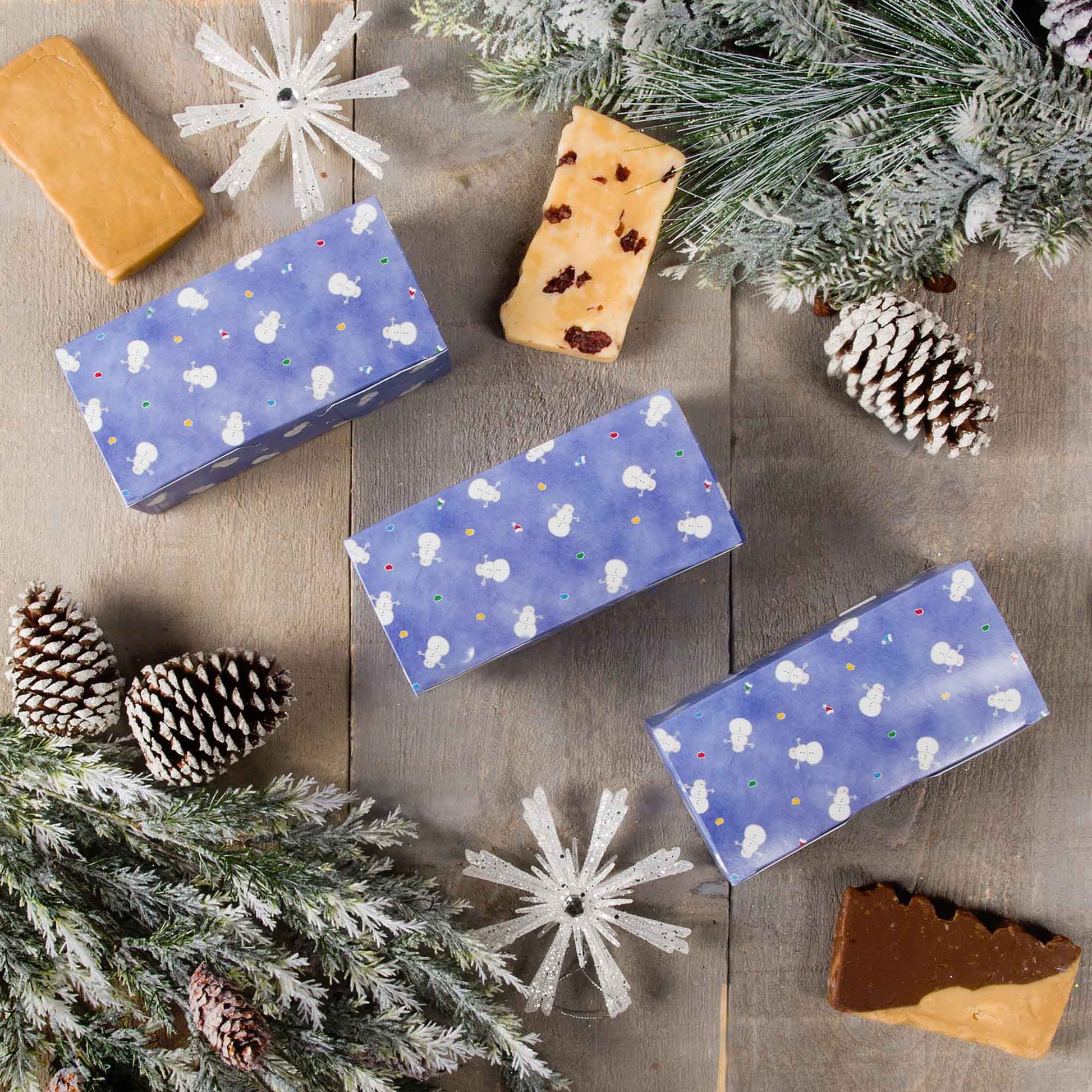 Blue snowman holiday candy boxes arranged on rustic table with winter greenery, pinecones, and assorted confections