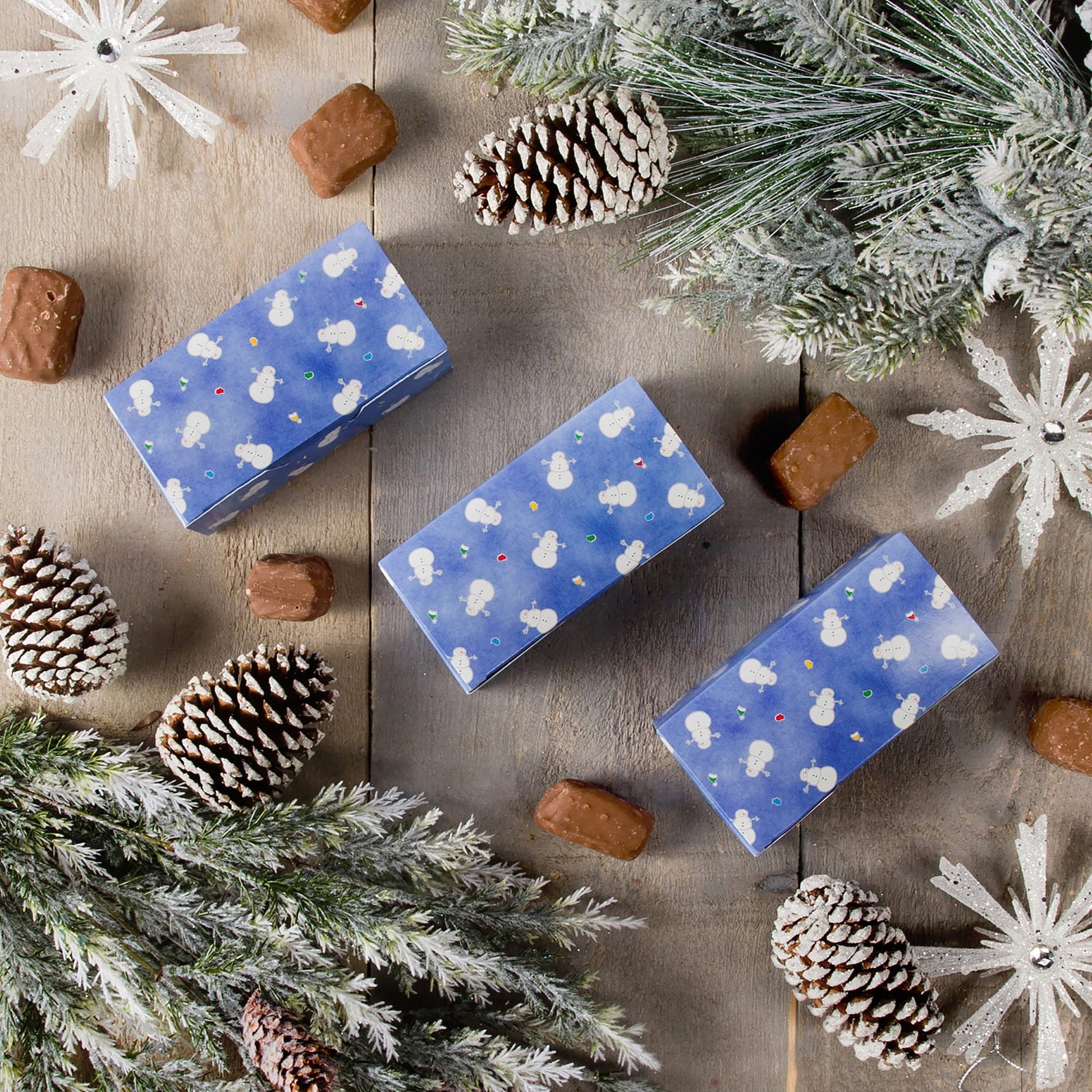 Three blue Snowman Avalanche candy boxes on a rustic tabletop with frosted pine branches and pinecones, surrounded by chocolates for a holiday gift display.