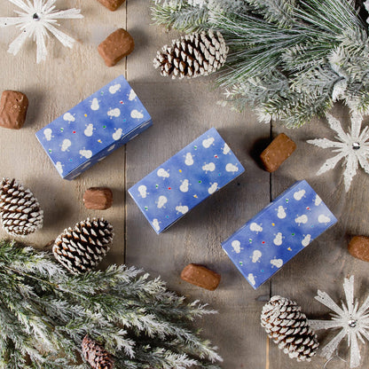 Three blue Snowman Avalanche candy boxes on a rustic tabletop with frosted pine branches and pinecones, surrounded by chocolates for a holiday gift display.