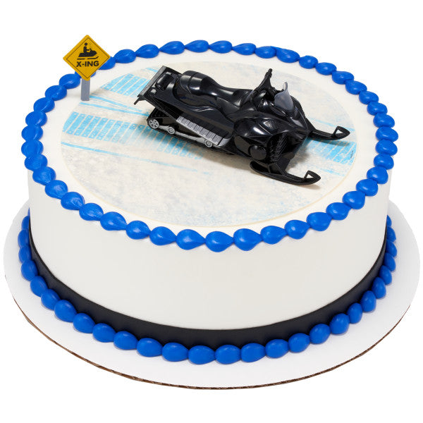 Round cake with white icing and bright blue bead border, topped with a black snowmobile figurine and a yellow snowmobile crossing sign.