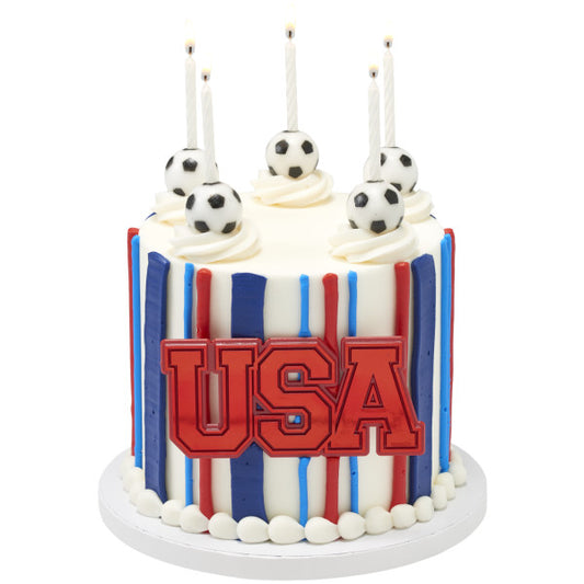 Tall red, white, and blue striped cake with USA plaque and soccer ball candle holders.