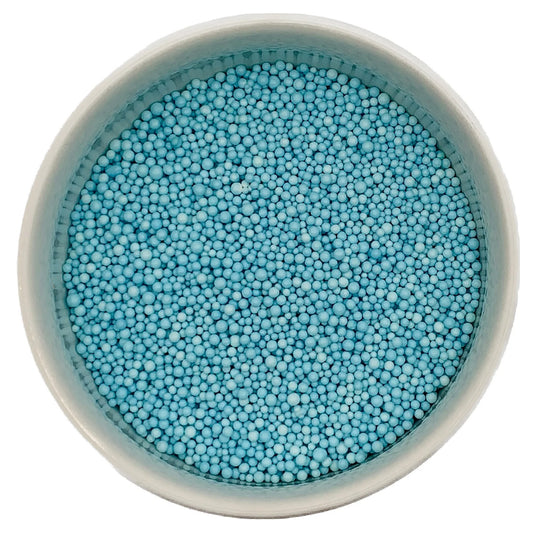Top-down view of a white bowl filled with soft blue non-pareils, showing tiny round sprinkle beads in a pale pastel blue shade.
