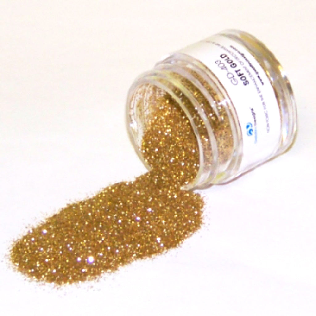 Small clear jar tipped on its side spilling soft gold glitter dust onto a white surface, with fine shimmering metallic particles visible throughout.