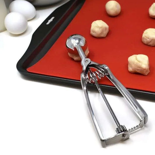 Cookie scoop resting on a red silicone-lined baking sheet with evenly portioned raw dough balls in the background.