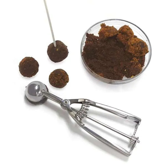 Stainless steel cookie scoop next to a bowl of cake pop mixture and several formed cake balls, one on a stick, demonstrating portioning use.