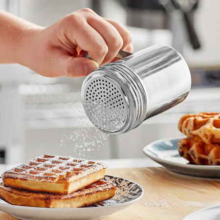 Plate of stacked waffles next to another plate with powdered sugar-dusted waffles, shown beside a stainless steel shaker on a wooden surface.