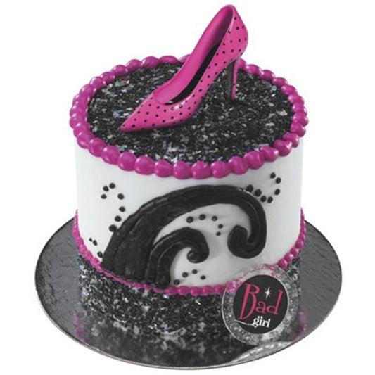 White frosted cake with black scroll piping and pink bead border, topped with a hot pink polka dot stiletto heel decoration on a black crumb-like surface.