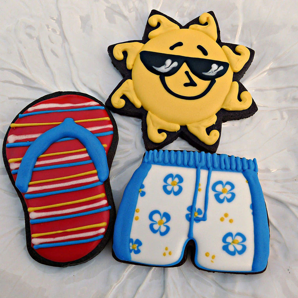 A set of decorated summer-themed cookies, including a sun wearing sunglasses, striped flip-flops, and blue floral swim trunks.