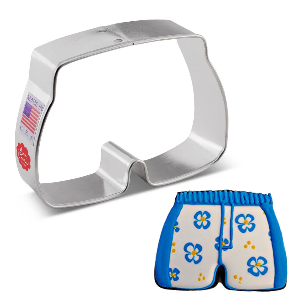 Metal swim trunks cookie cutter next to a decorated sugar cookie shaped like swim trunks, featuring blue trim and a floral pattern.