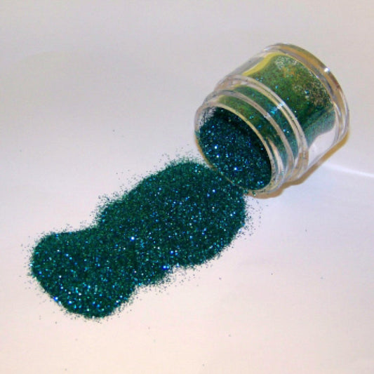 Small jar of teal glitter dust tipped on its side with teal glitter spilled onto a white surface.