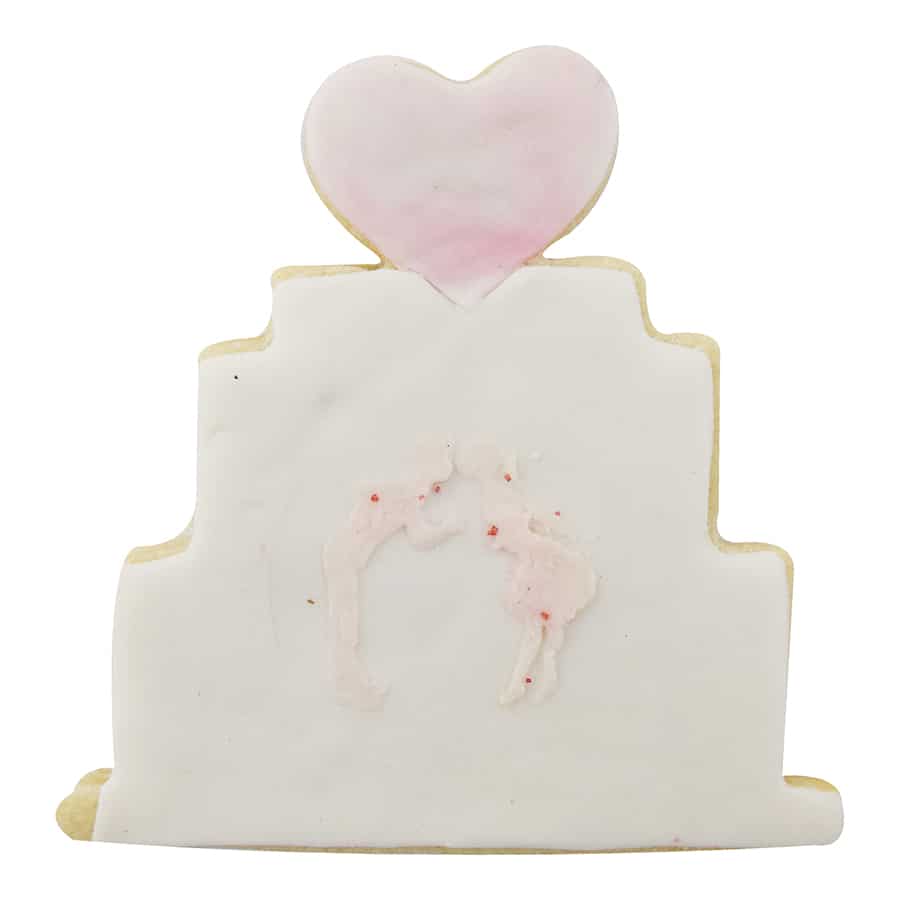 Decorated wedding cake-shaped sugar cookie with white icing, a pink heart topper, and a subtle embossed design of a kissing couple.