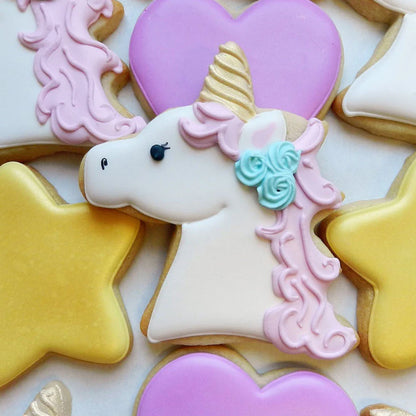 Unicorn sugar cookie with white icing, a pink mane, a golden horn, and blue floral details, surrounded by heart and star cookies.