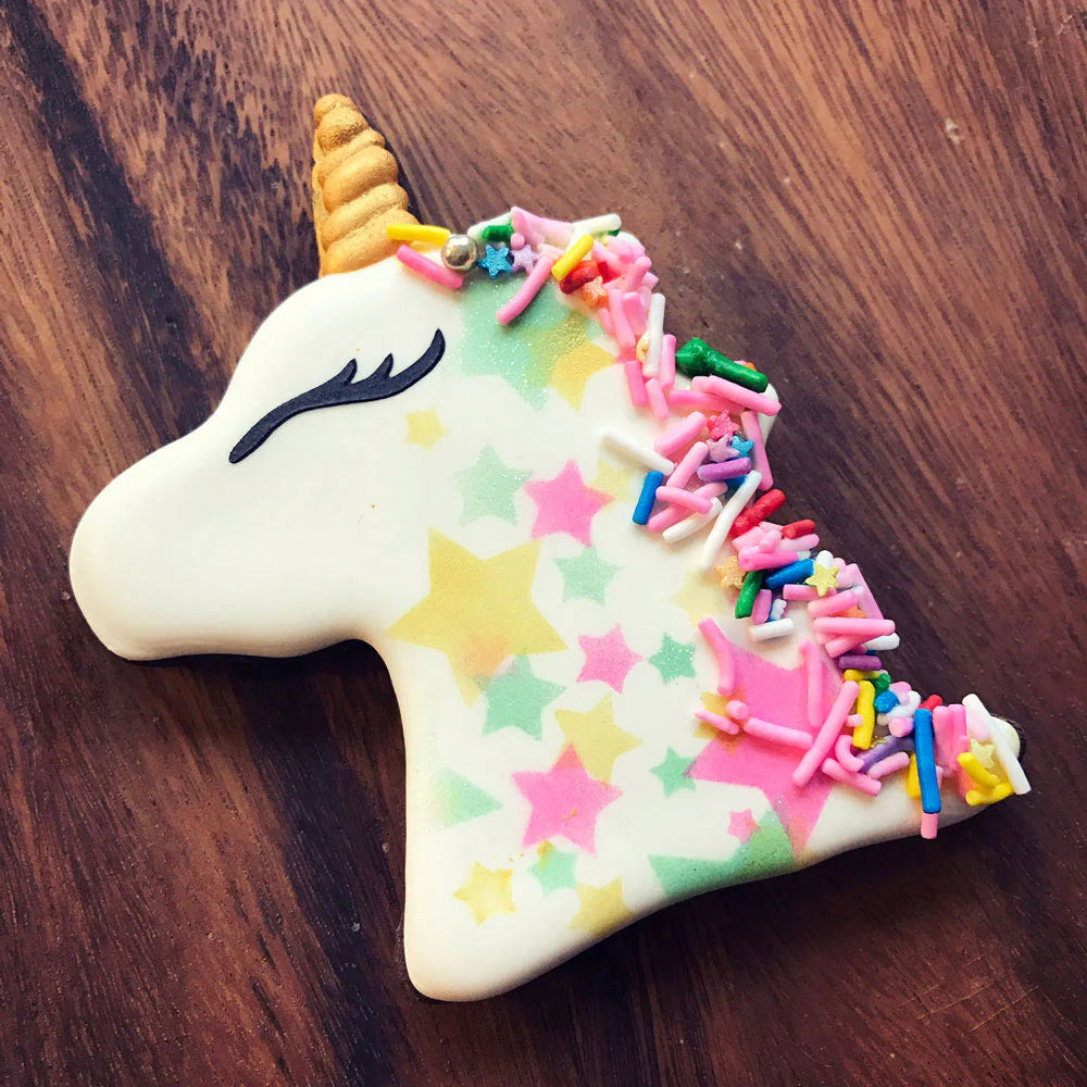 Decorated unicorn-shaped sugar cookie with white icing, a golden horn, colorful sprinkles, and pastel star patterns.