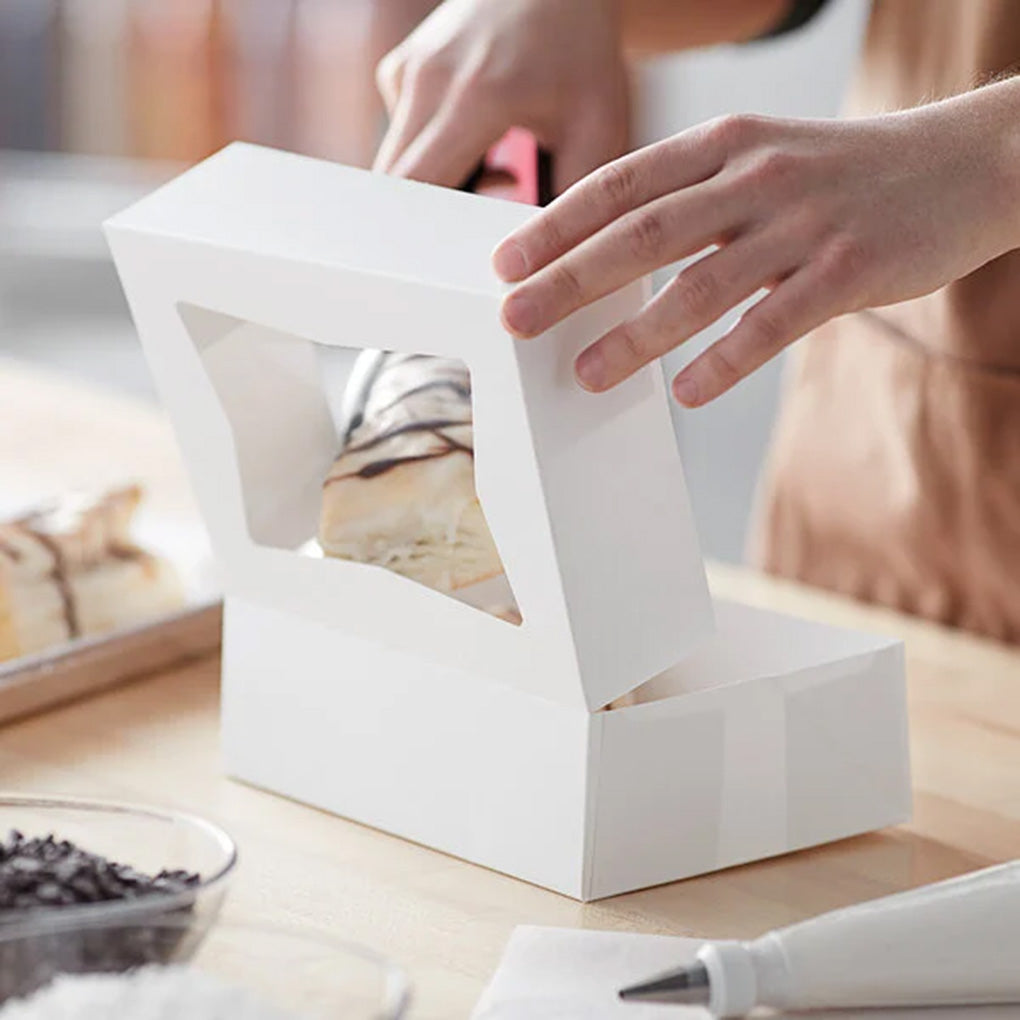 Hands closing a white bakery box with a window over a drizzled pastry inside, on a kitchen work surface with baking tools nearby.
