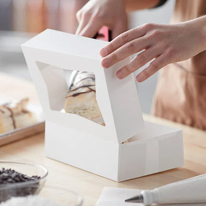 Hands closing a white bakery box with a window over a drizzled pastry inside, on a kitchen work surface with baking tools nearby.