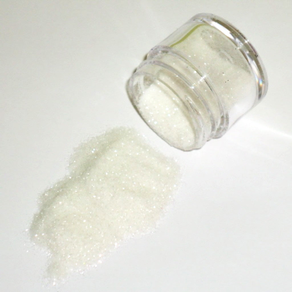Small clear jar tipped on its side spilling white glitter dust onto a white surface, with fine shimmering particles visible throughout.