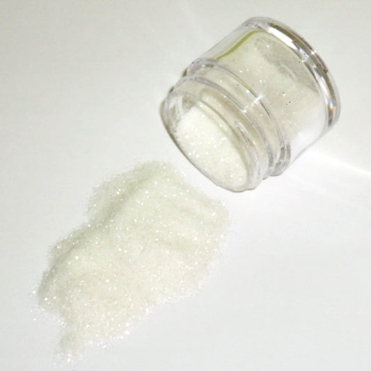 Small clear jar tipped on its side spilling white glitter dust onto a white surface, with fine shimmering particles visible throughout.
