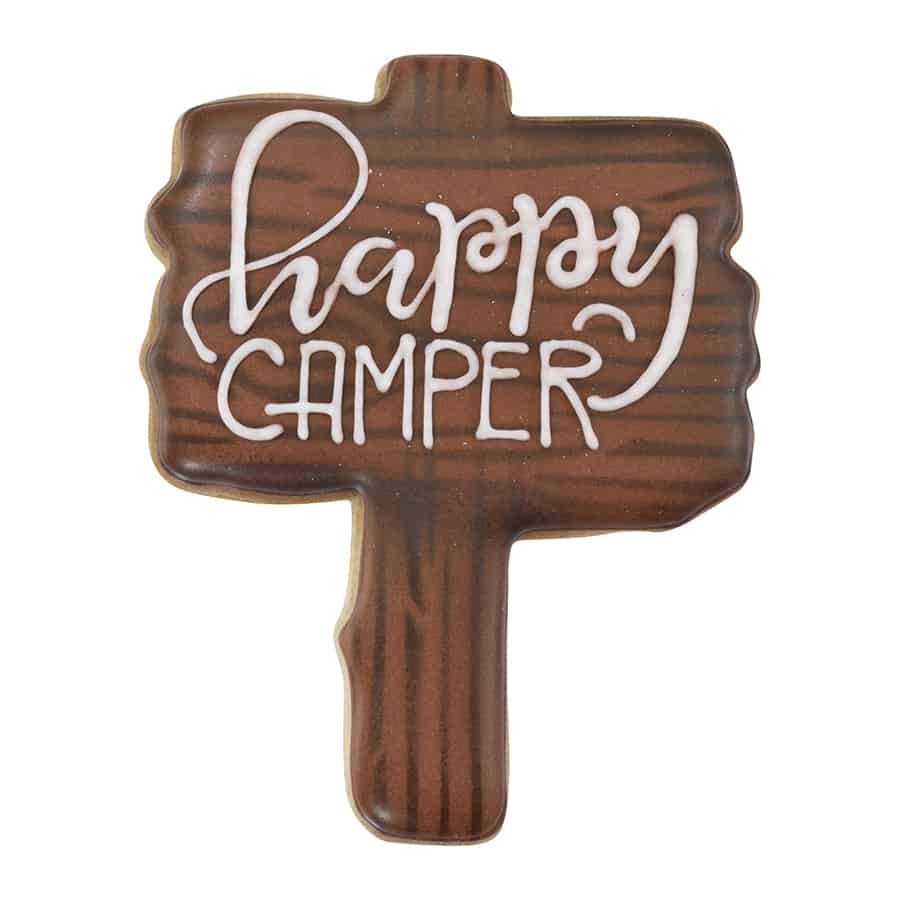 A decorated wood sign cookie with brown icing and the words "Happy Camper" written in white.