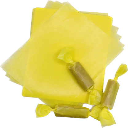 Three rectangular caramel pieces individually packaged in yellow wax candy wrappers with twisted ends, displayed beside a stack of 4" x 5" caramel wrapper sheets.